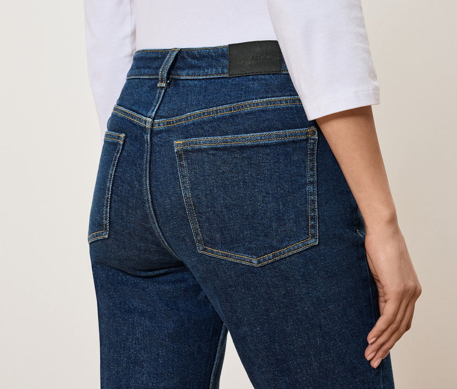 Hátulnézet egy nőről, aki magas derekú, kék farmernadrágot és fehér pólót visel.