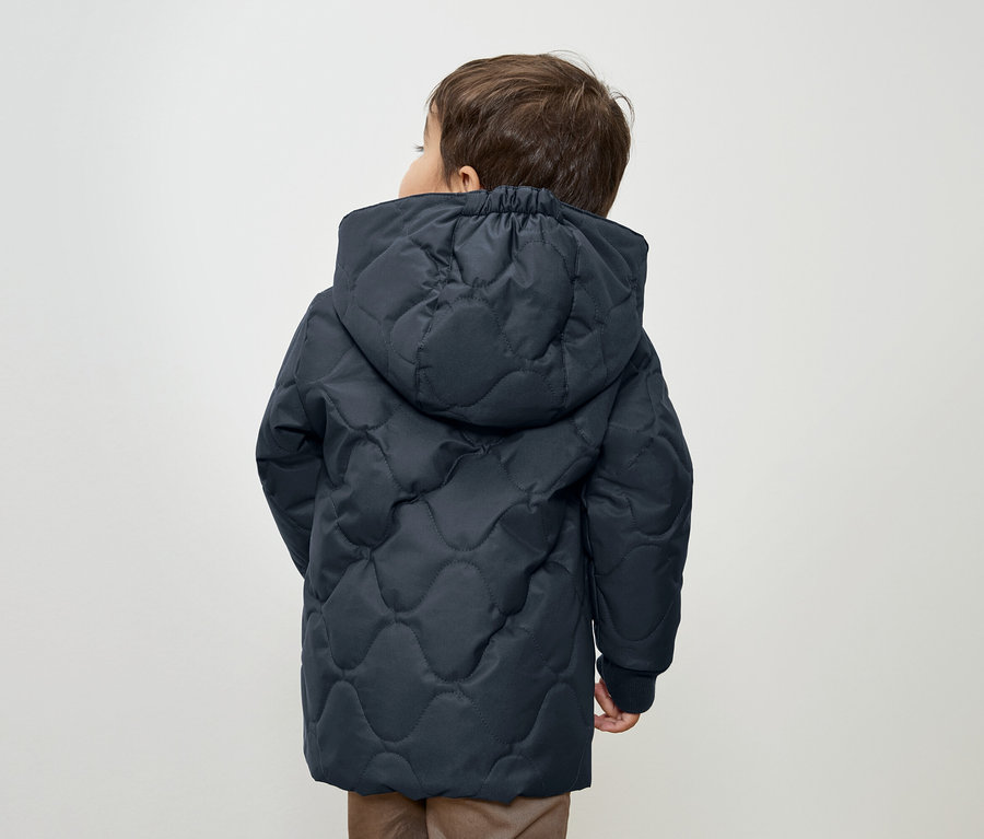 Fiú sötétkék, steppelt, kapucnis kabátban, háttal a kamerának.