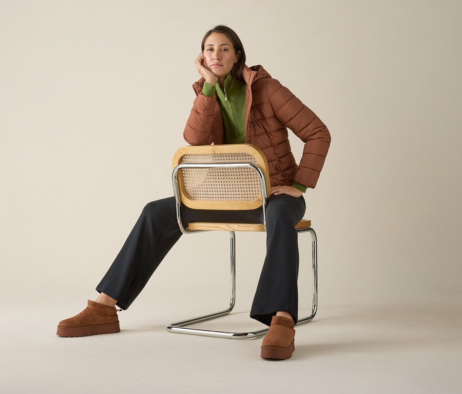 Nő ül egy széken kötött magasnyakú pulóverben, kabátban és cipőben.