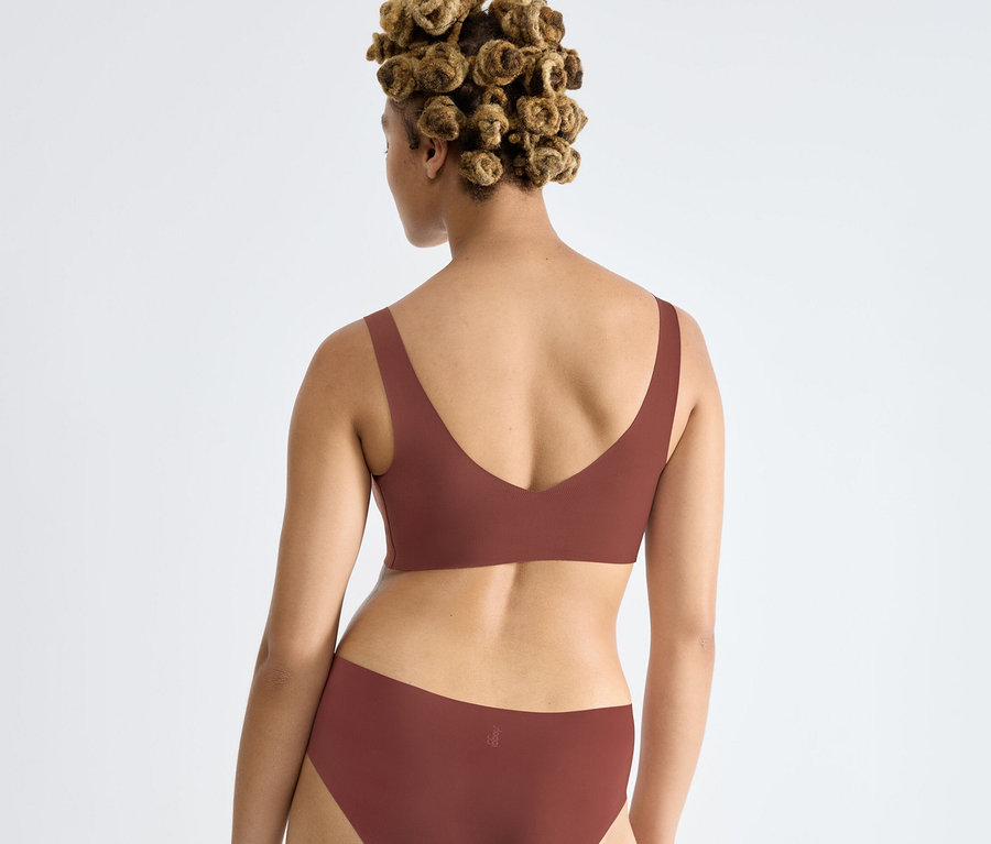 Nő hátulról, barna sloggi ZERO Feel 2.0 Bralette melltartóban és hozzá illő alsóneműben, göndör hajjal, kis kontyokba kötve.