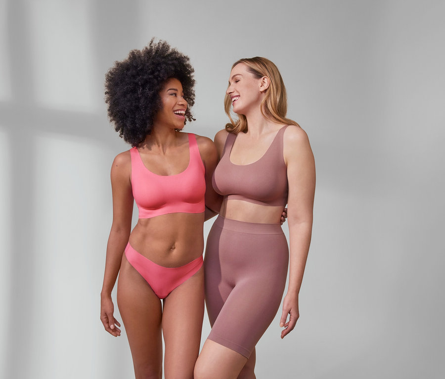 Két nő nevetve öleli egymást. Egyikük rózsaszín Női komfort bustier melltartót és 5 női tangát, Magic Cut-ot visel. A másik Női seamless rövidnadrágot és egy felsőt visel.