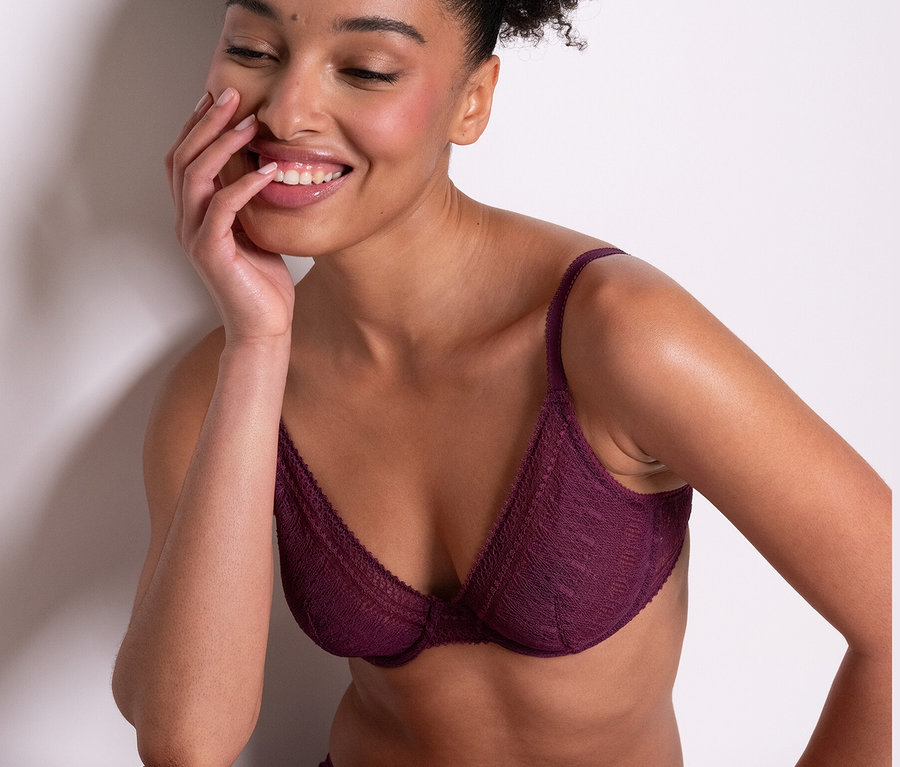 Egy mosolygó nő ül, lila Passionata merevítős melltartót »Leila« és hozzáillő alsót visel.
