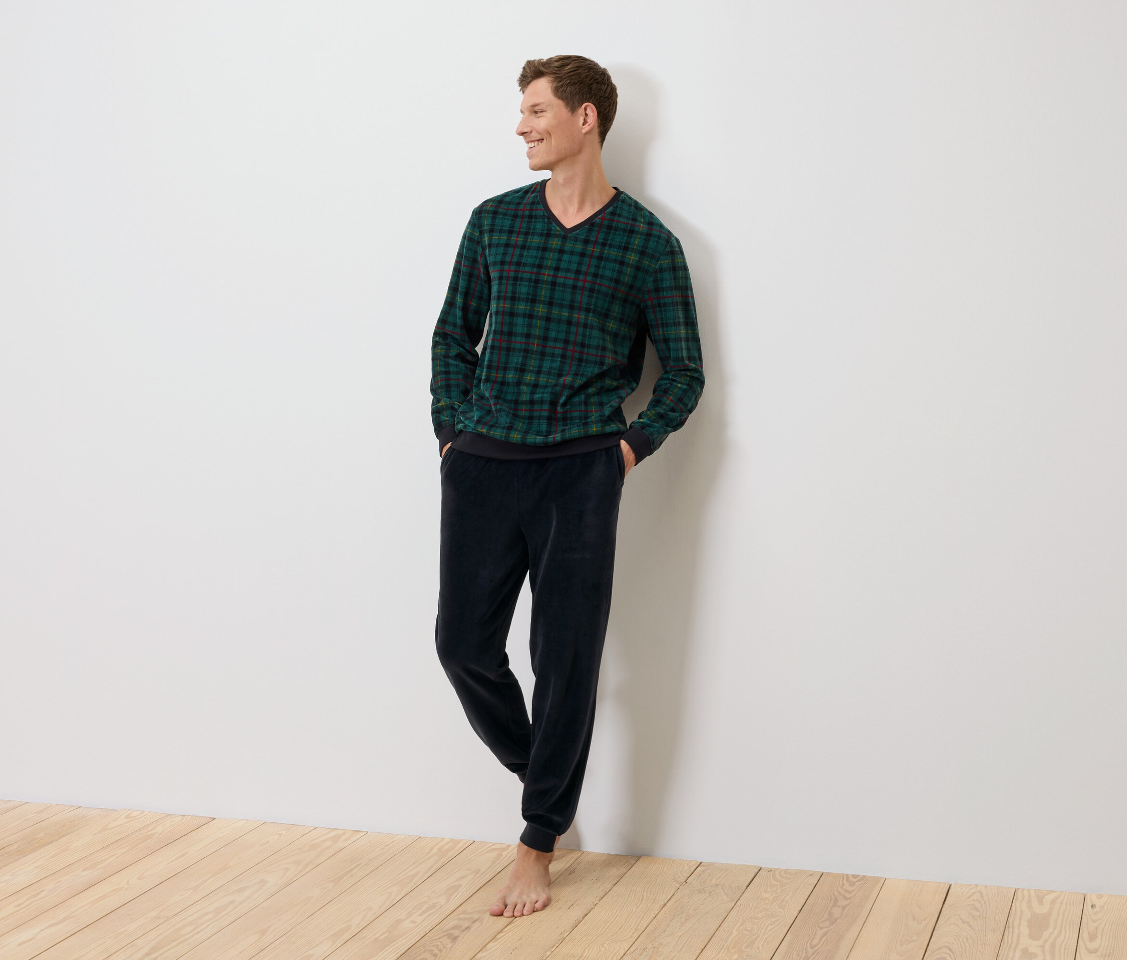 Egy férfi áll a falnak támaszkodva, zöld kockás pulóvert és sötétkék bársonynadrágot visel.