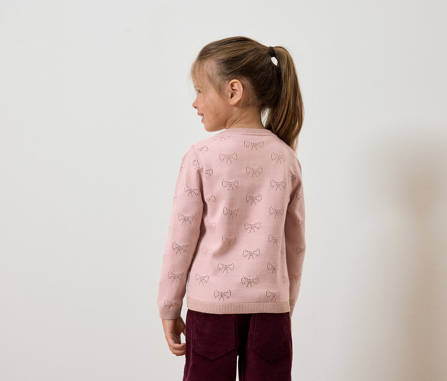 Egy kislány copfban, rózsaszín, masnis pulóverben és bordó nadrágban oldalról.