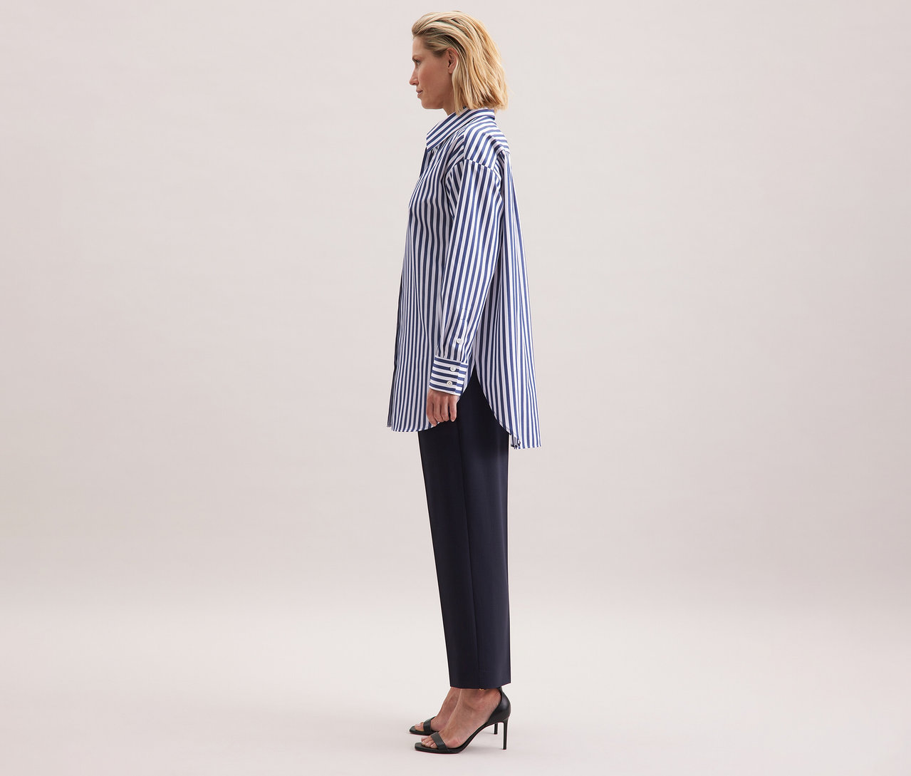 Nő visel egy csíkos Seidensticker túlméretes hosszú ingblúzt, sötét nadrágot és fekete szandált.