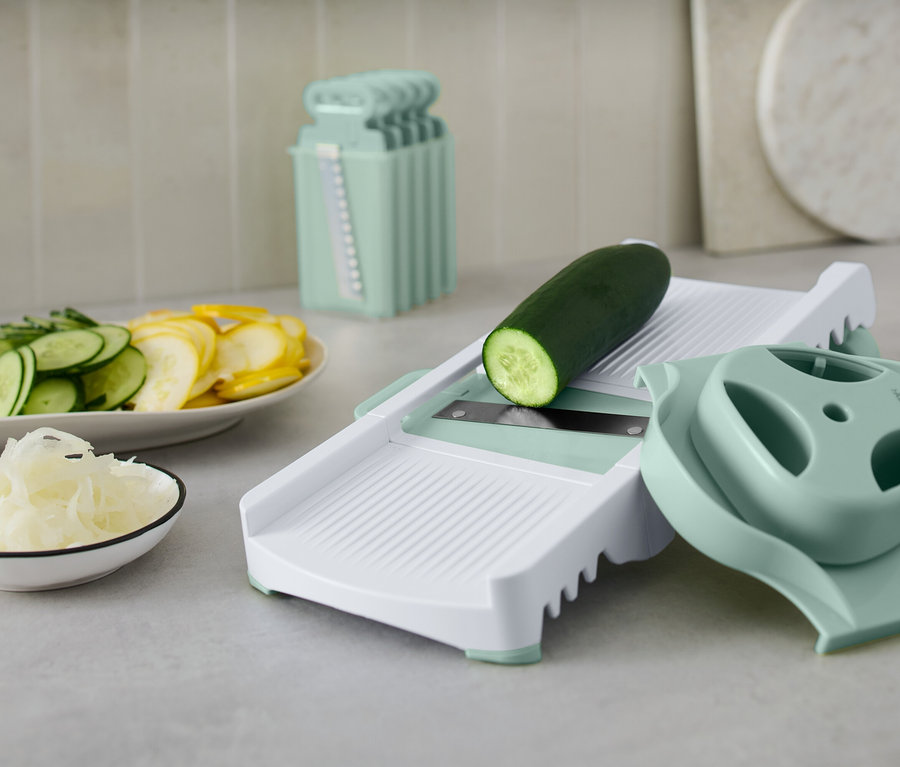 Zöld uborka egy fehér zöldségszeletelőn tartozékokkal. Egy tányéron szeletelt uborka és sárga tök található.