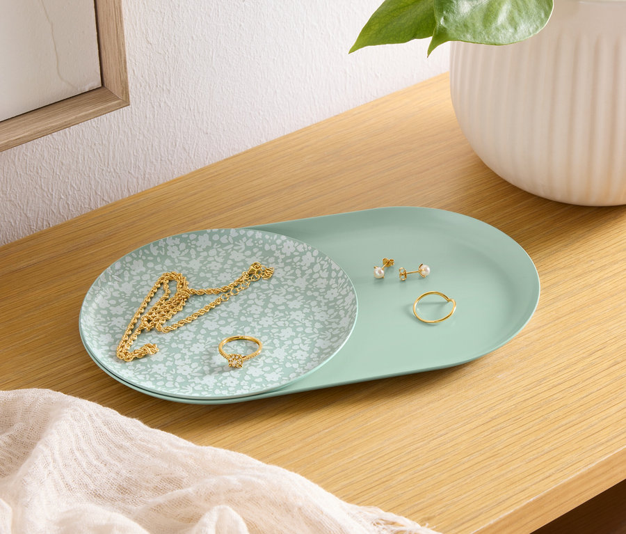 Egy fa polcon egy zöld tálca található, rajta egy arany nyaklánc, egy gyűrű és fülbevalók. A tálca mellett egy fehér váza áll.
