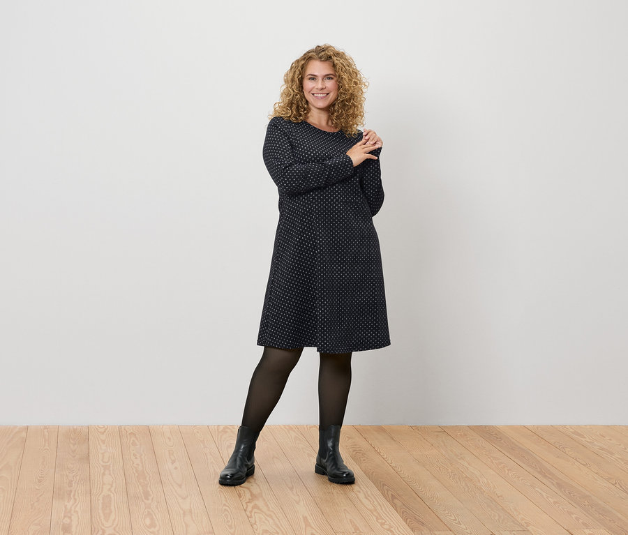 Egy nő pózol egy sötétkék, pöttyös ruhában, fekete harisnyában és csizmában.