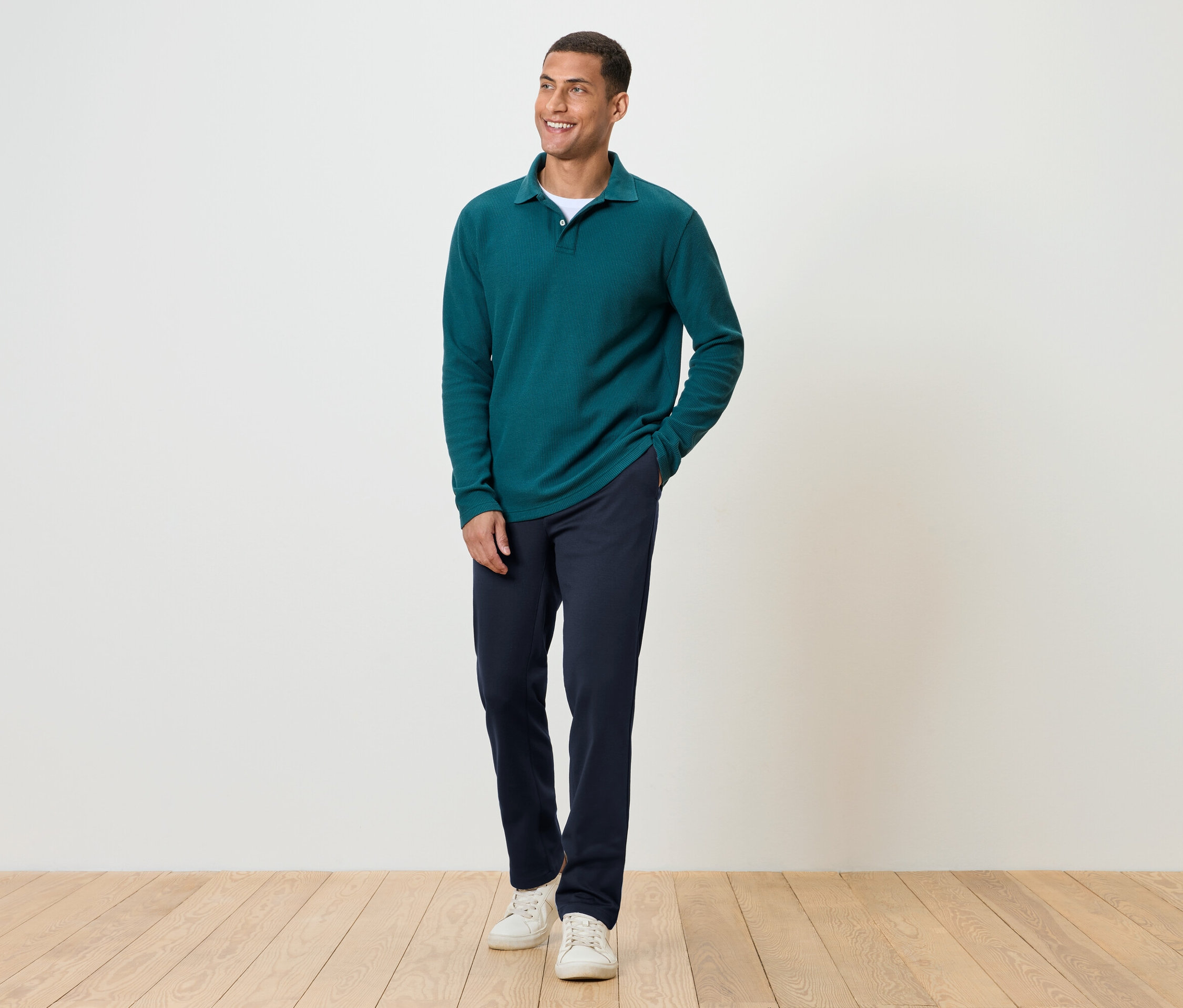 Egy férfi áll egy fapadlón, sötétkék nadrágot, zöld, hosszú ujjú pólót és fehér tornacipőt visel.