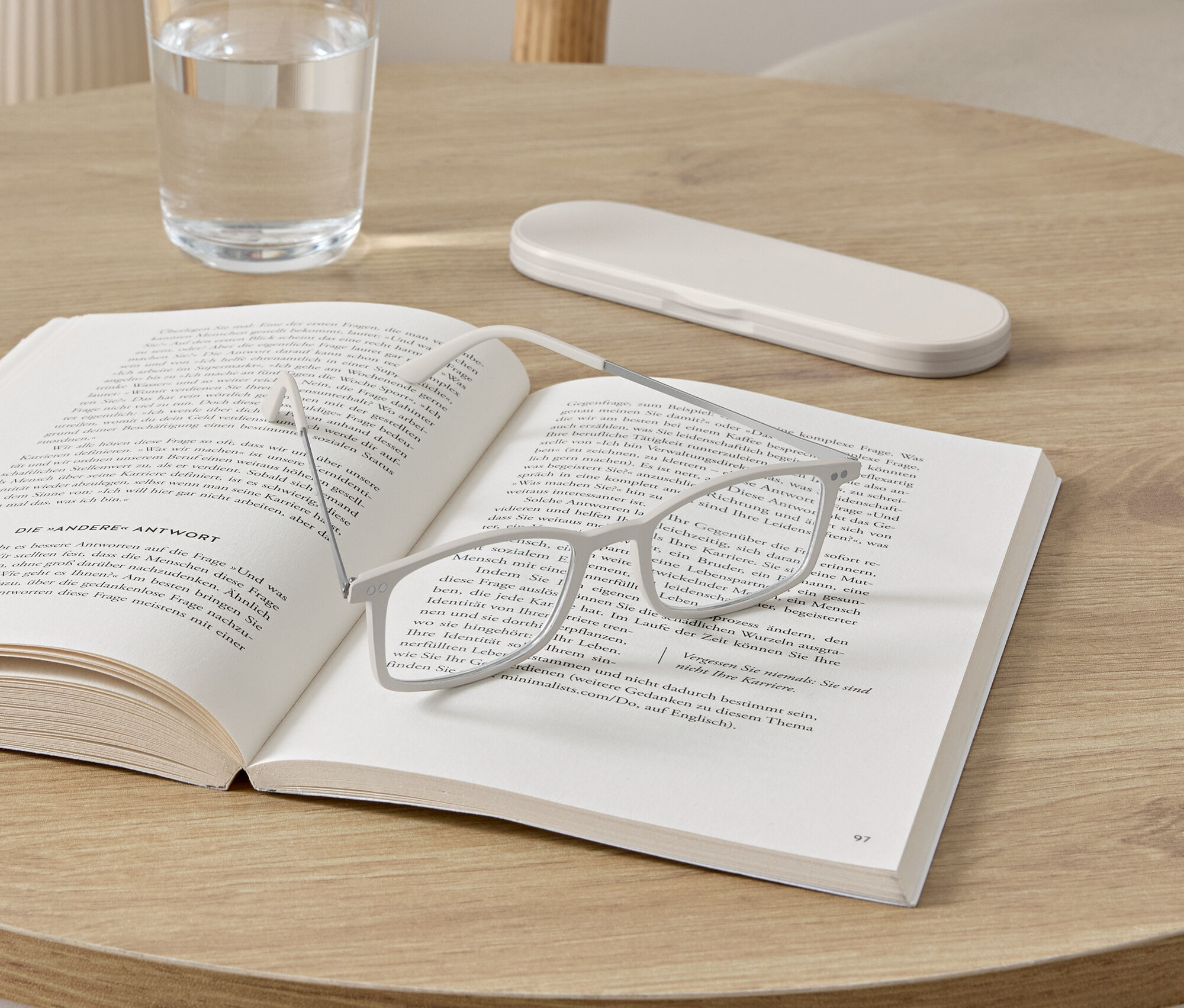 Egy szemüveg fekszik egy nyitott könyvön egy kerek asztalon. A könyv mellett egy pohár víz és egy szemüvegtok található.