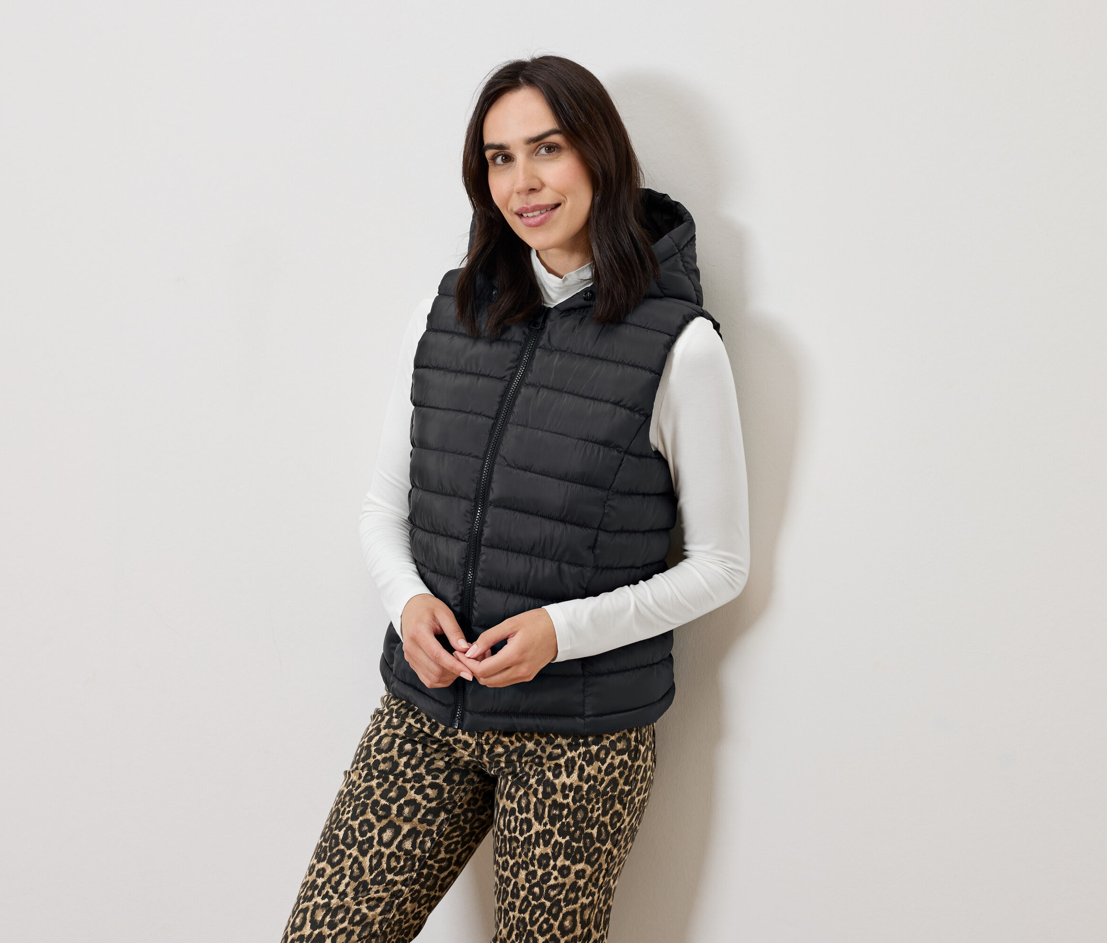 Nő pózol fekete, steppelt, kapucnis mellényben, fehér garbóban és állatmintás nadrágban.