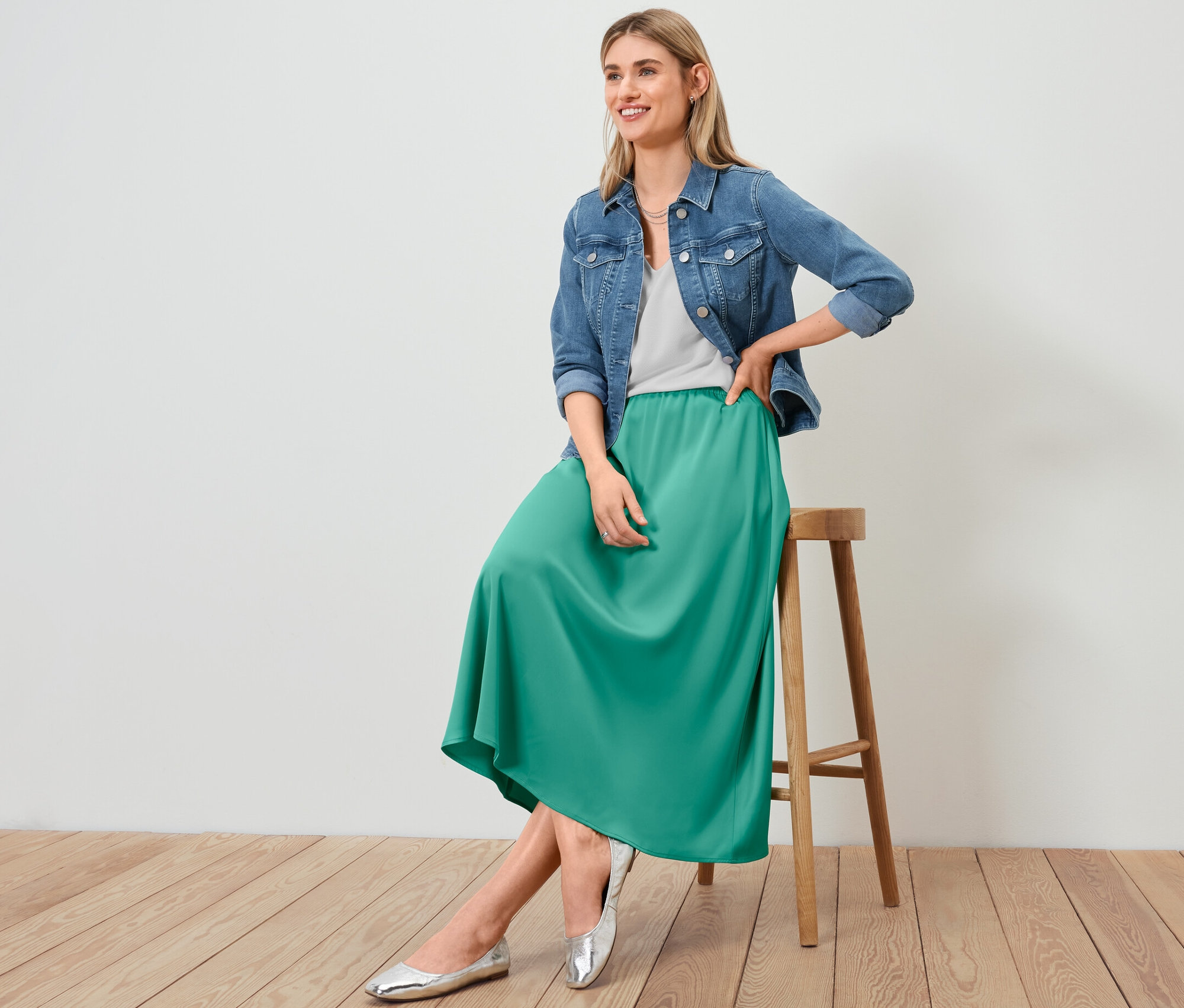 Nő ül egy széken farmerdzsekiben, szaténszoknyában és fehér, strukturált felsőben.