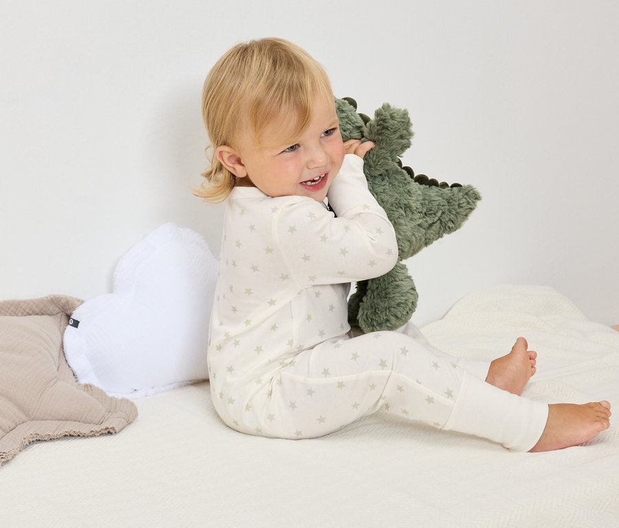Egy szőke hajú gyermek csillagos, fehér pizsamában ül egy fehér takarón, és egy zöld dinoszaurusz plüssállatot ölel.