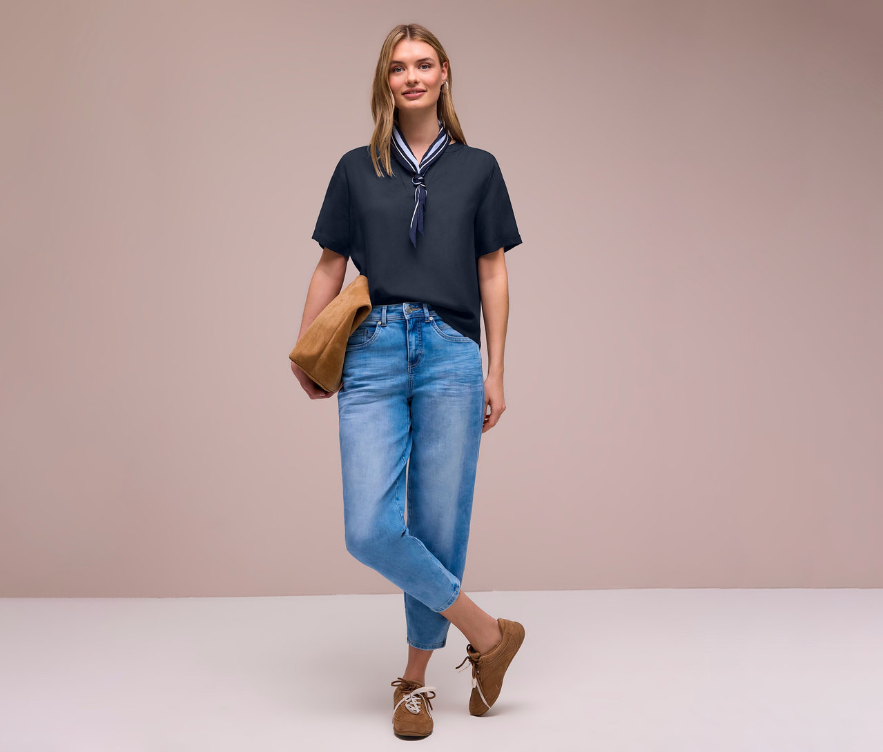 Farmerben és barna cipőben, táskával egy nő Street One blúzt visel sállal.