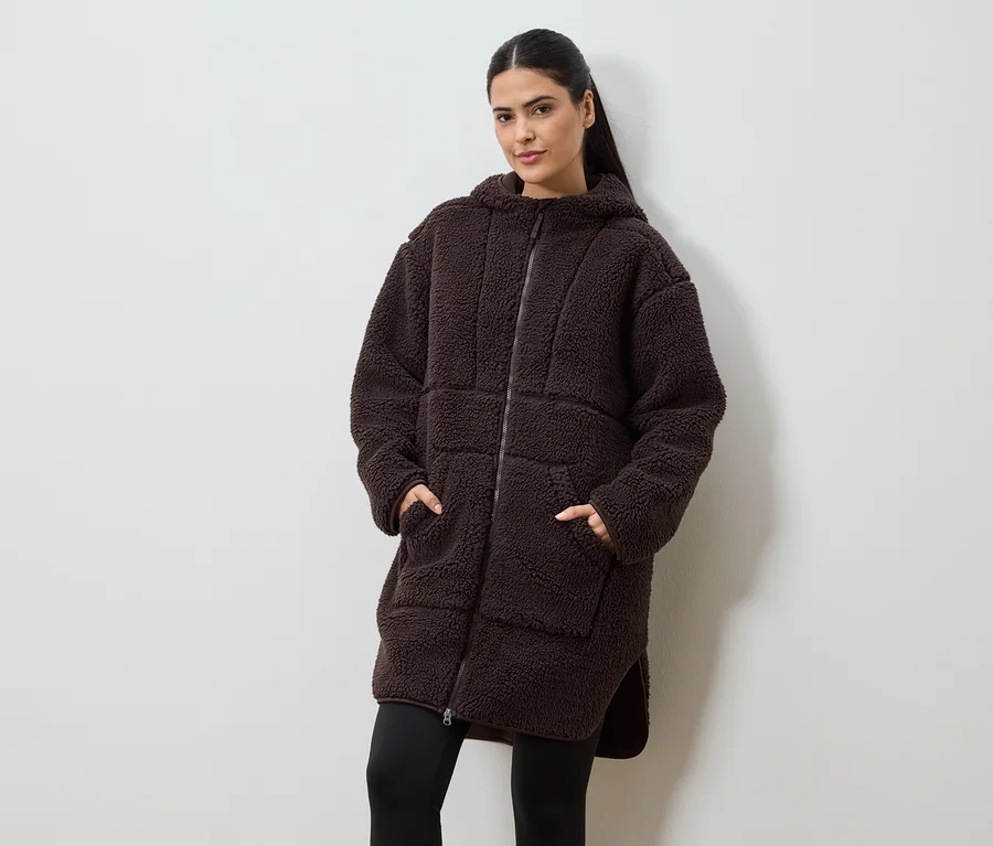 Zsebében a kezével egy nő hosszú, barna, kapucnis, cipzáras, teddykabátot visel fekete leggings fölött.
