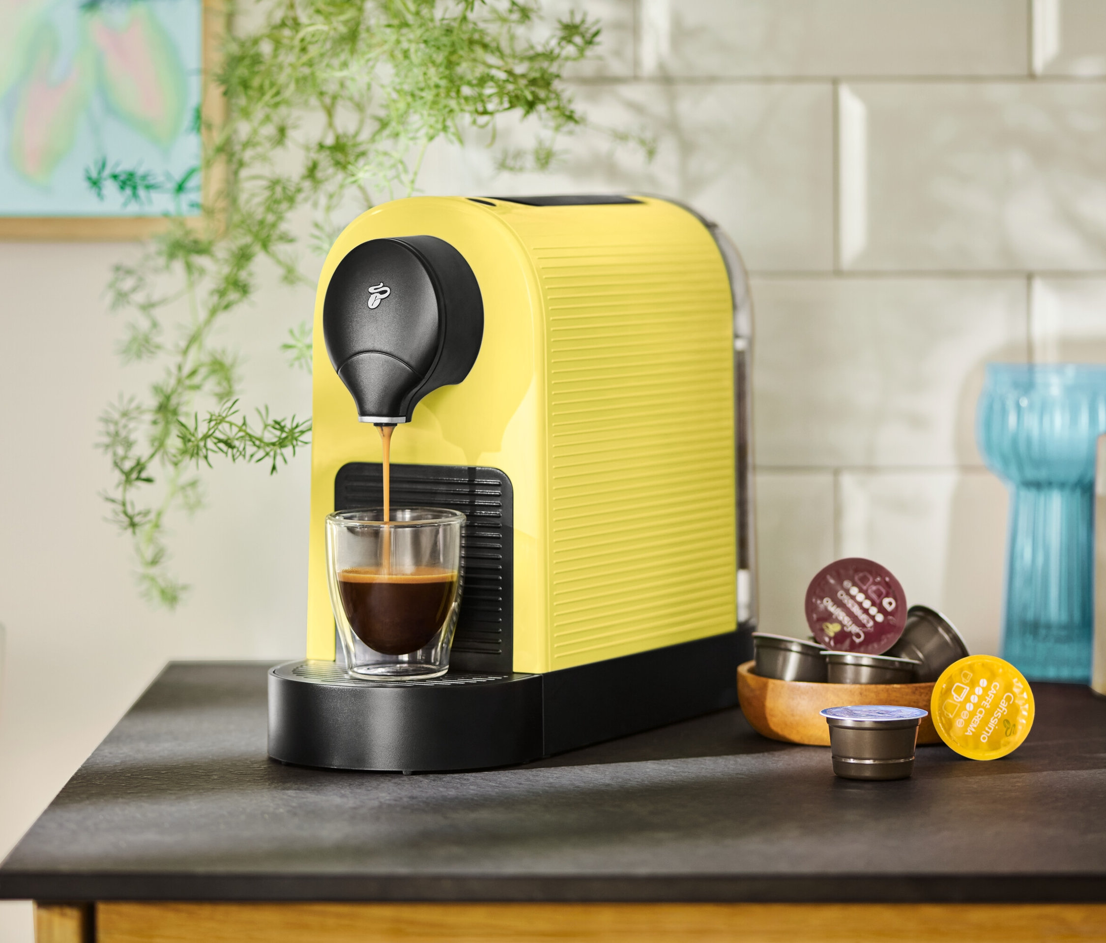 Egy sárga kávéfőző kávéval tölt egy csészét, mellette kapszulák vannak.