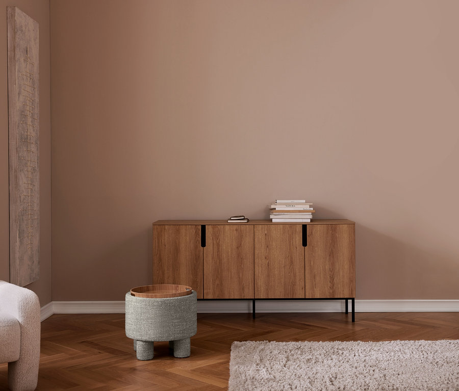 Modern nappali bézs falakkal és halszálka parkettával. Fa tálalószekrény könyvekkel, szürke kárpitozott puff fa tálcával, szürke fotel, világos szőnyeg.