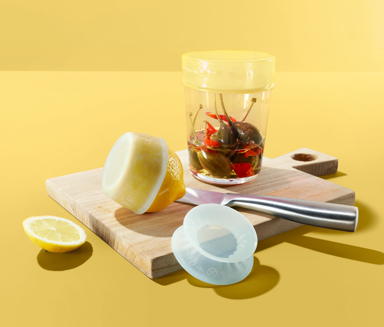 Sárga háttér előtt vágódeszka egy üveg chilipaprikával, egy citrommal és 3 db-os frissentartó szilikon fedél szettel.