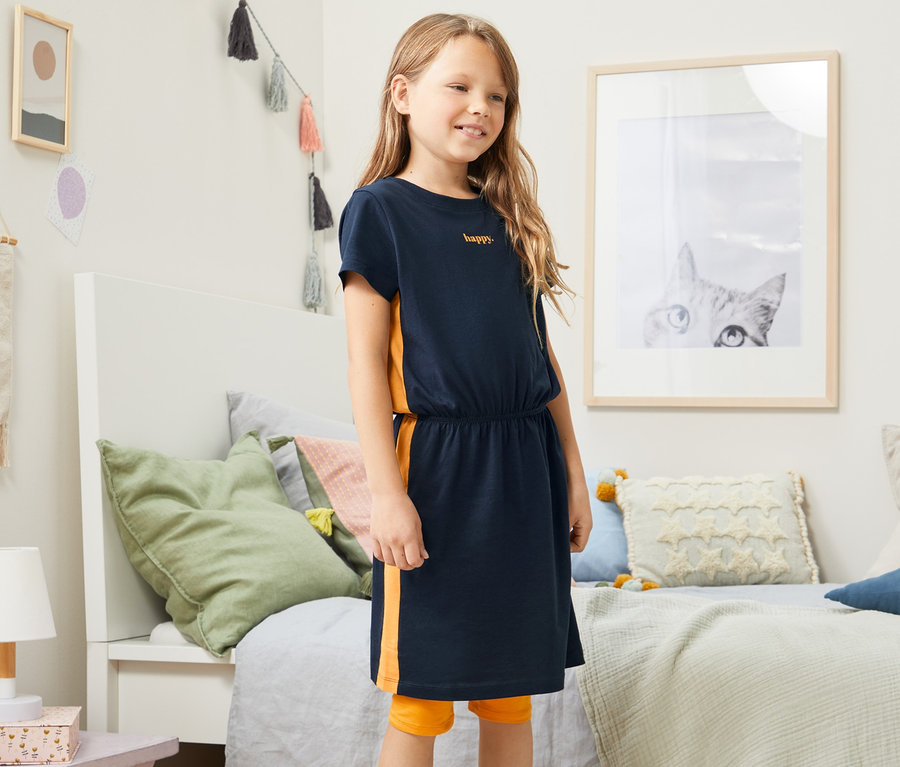 Egy lány pózol a hálószobában, kék dzsörzéruhát visel.