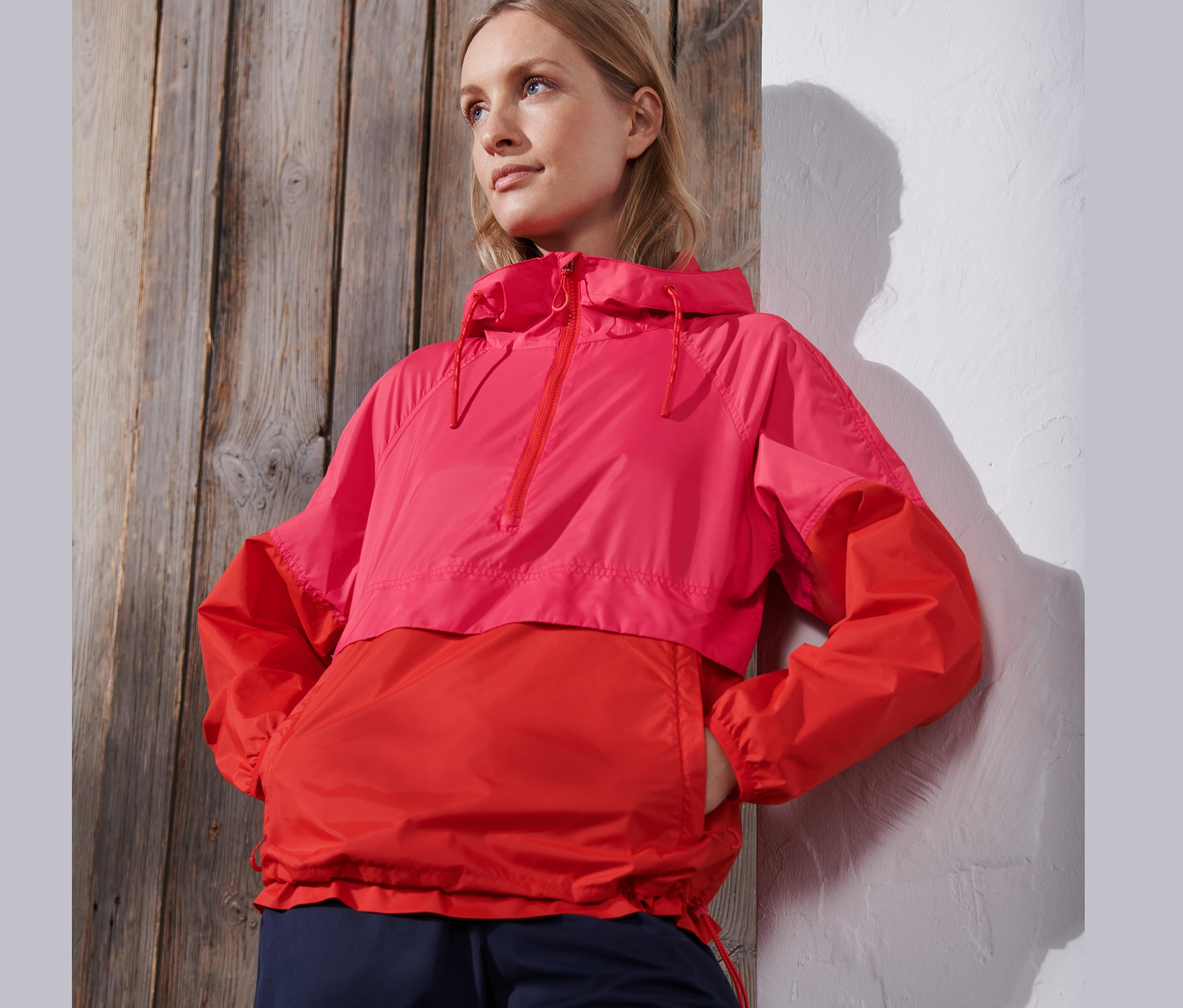 Egy falnak támaszkodó nő rózsaszín/piros női széldzsekit és sötétkék nadrágot visel.