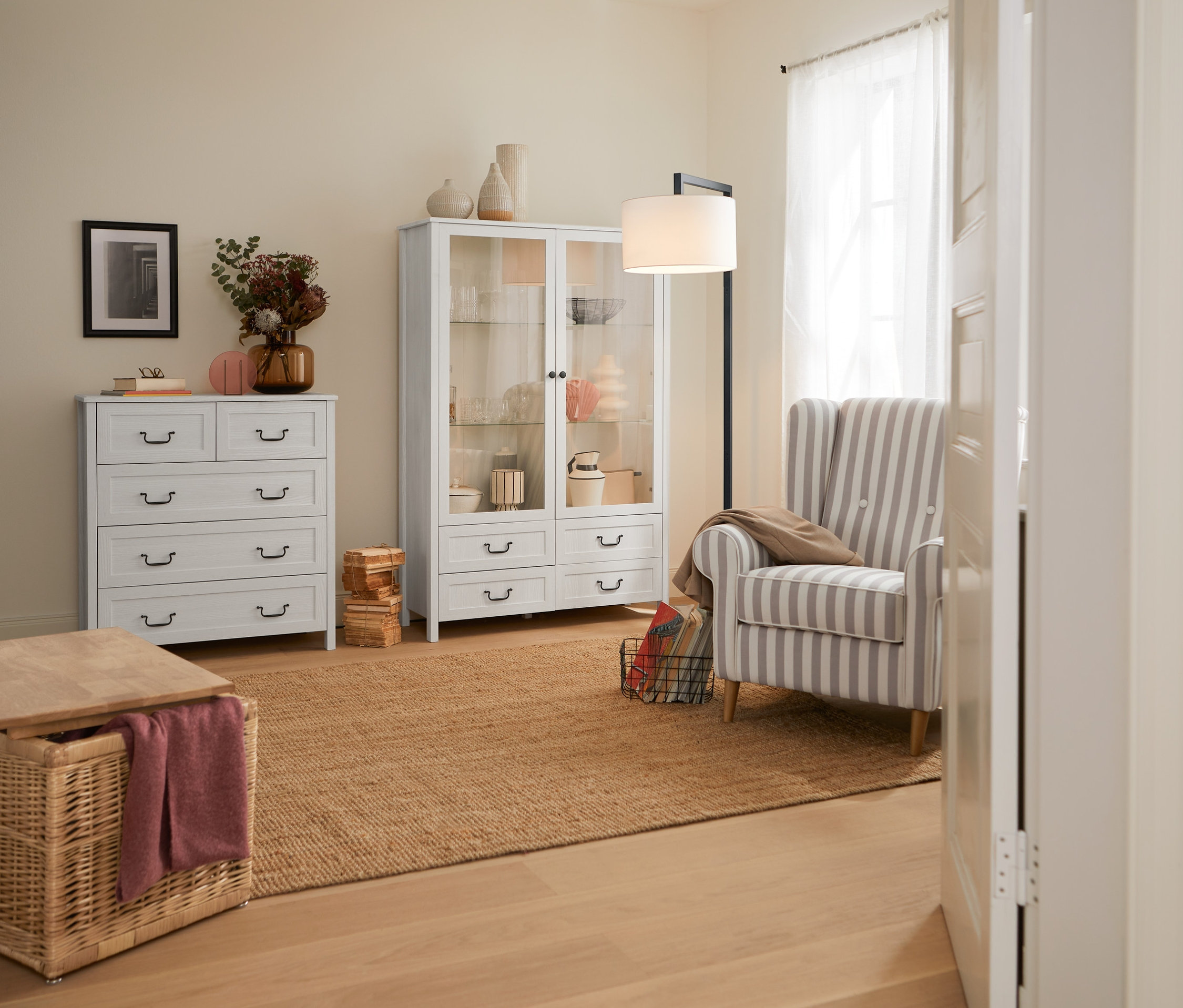 Belső tér rattan tárolóládával, fehér komóddal, 5 fiókkal, fehér vitrines szekrénnyel, 2 ajtóval, 4 fiókkal és fekete fém LED-es állólámpával.