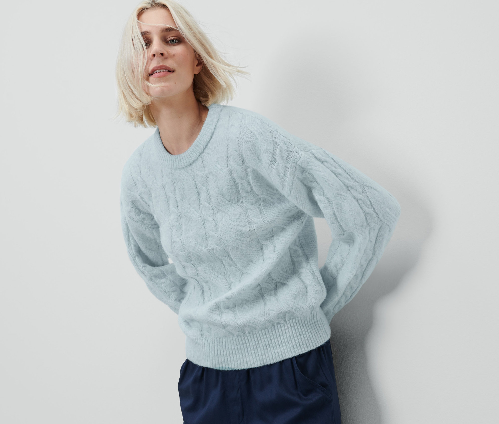 Nő pózol fehér háttér előtt, világoskék kötött pulóvert visel.