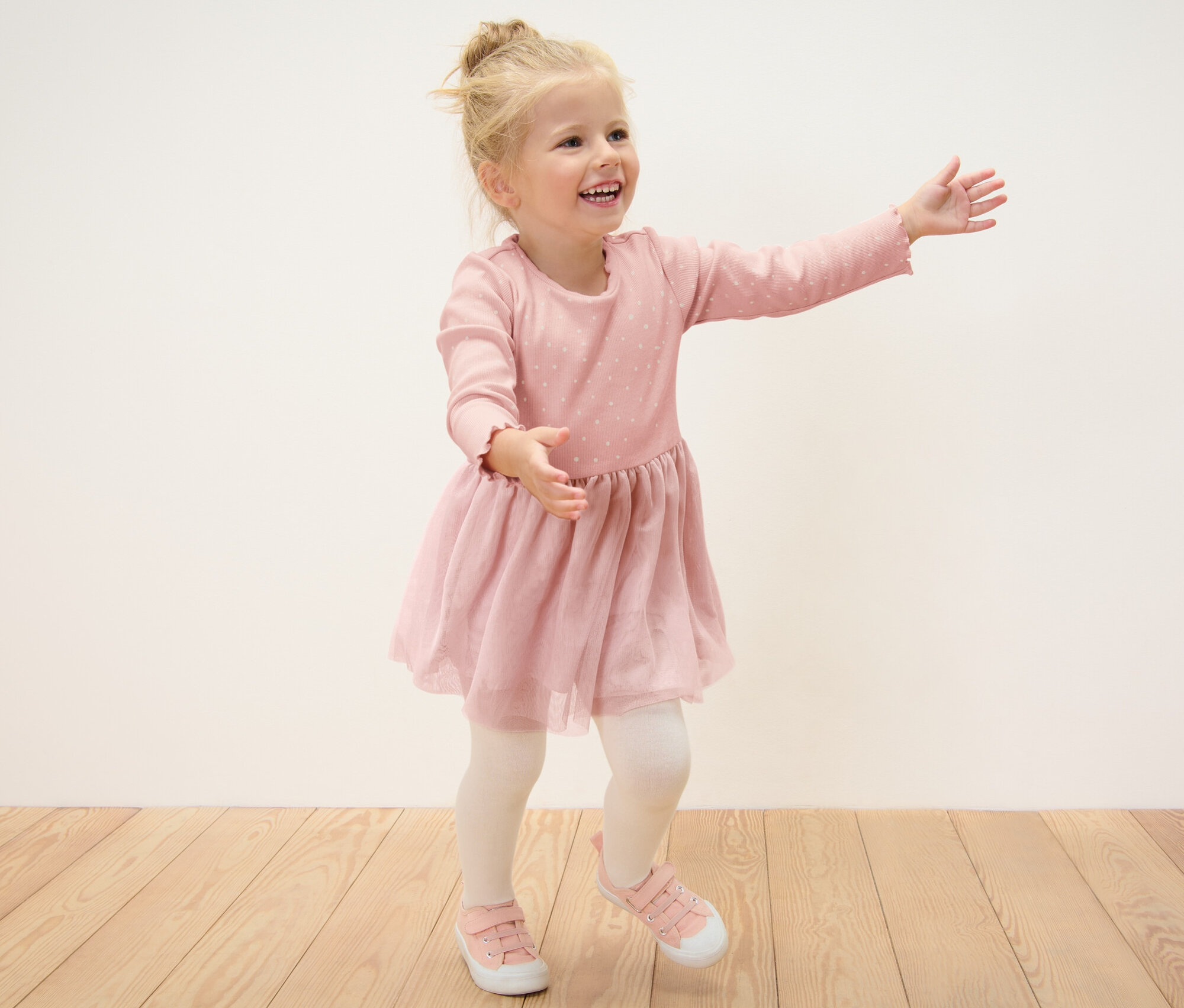 Egy mosolygós szőke kislány bordás ruhában pózol a fapadlón.