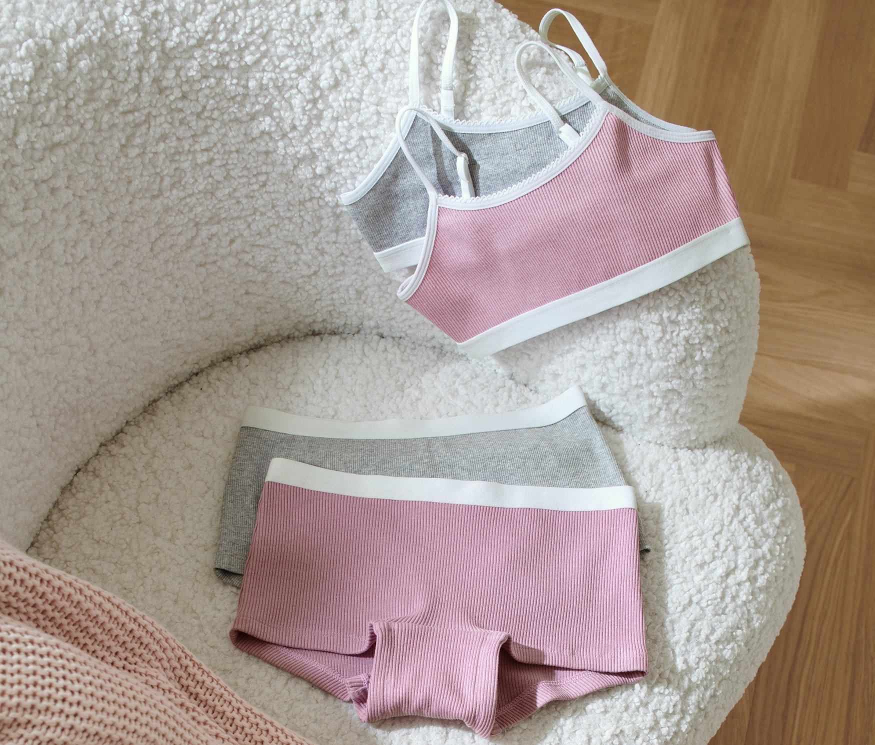 Egy lány fehérnemű szett egy fehér, bozontos széken fekszik egy rózsaszín kötött takaró mellett.