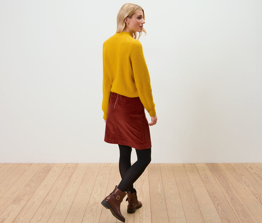 Szőke nő sárga pulóvert és női kordbársony szoknyát visel. Fapadlón áll.