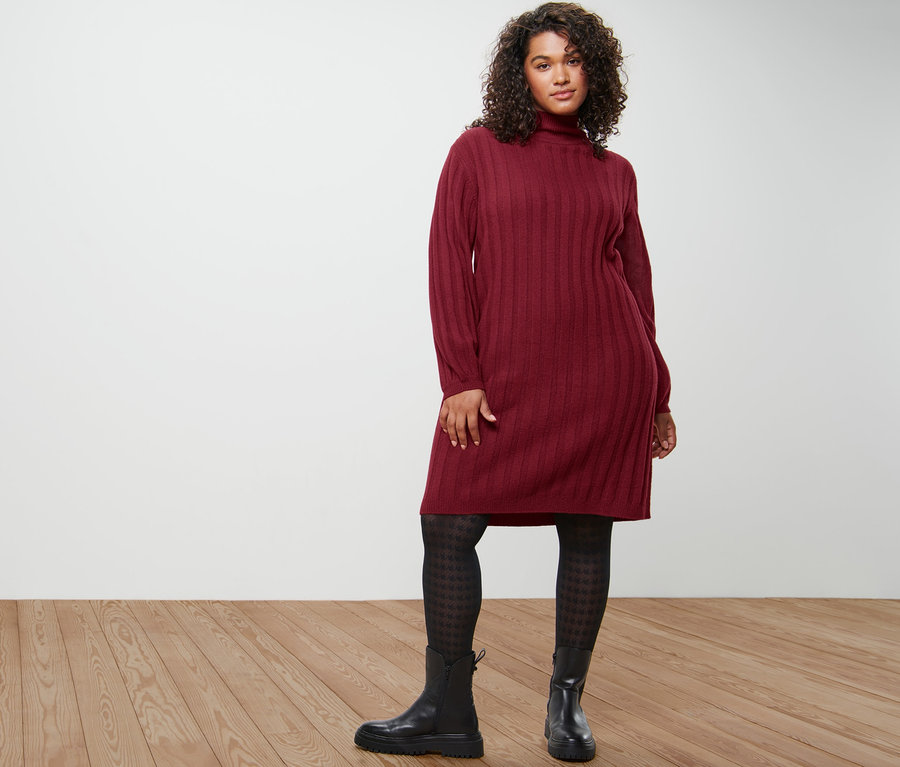 Nő pózol bordázott kötött ruhában, tyúklábmintás harisnyában és fekete bőrcsizmában.