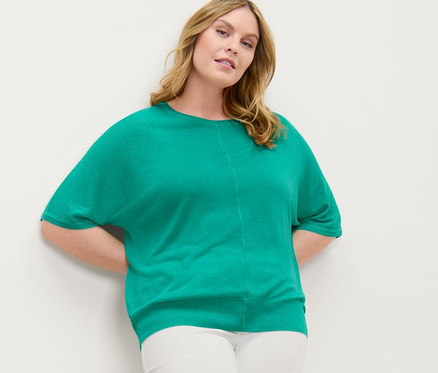 Nő pózol a falnak dőlve, finomkötésű felsőt és 7/8-os, lyukhímzéses tregginget visel.