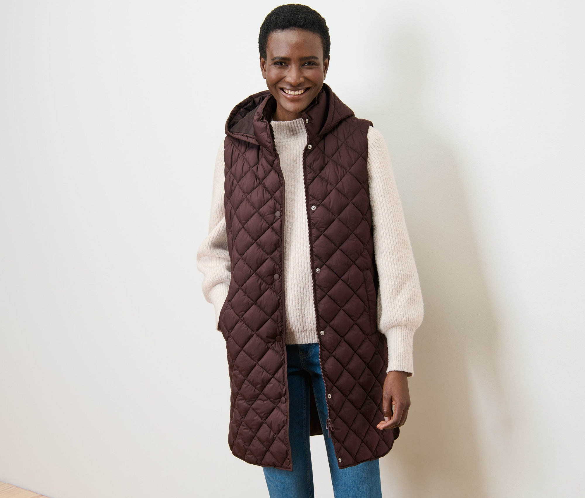 Mosolygó nő pózol egy sötétbarna, hosszú steppelt mellényben, kapucnival.