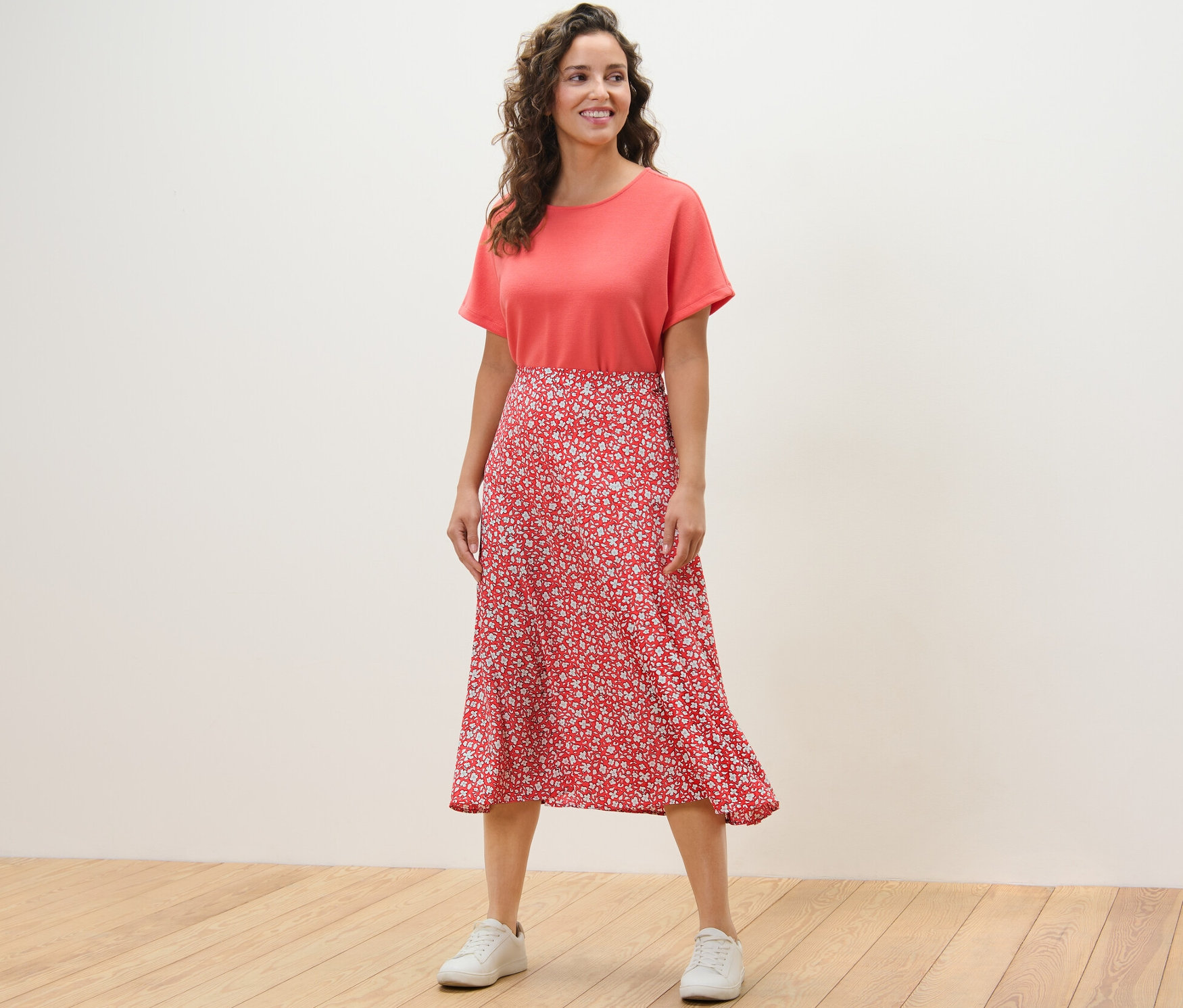 Nő áll a fapadlón, korall színű, mintás midiszoknyát visel.