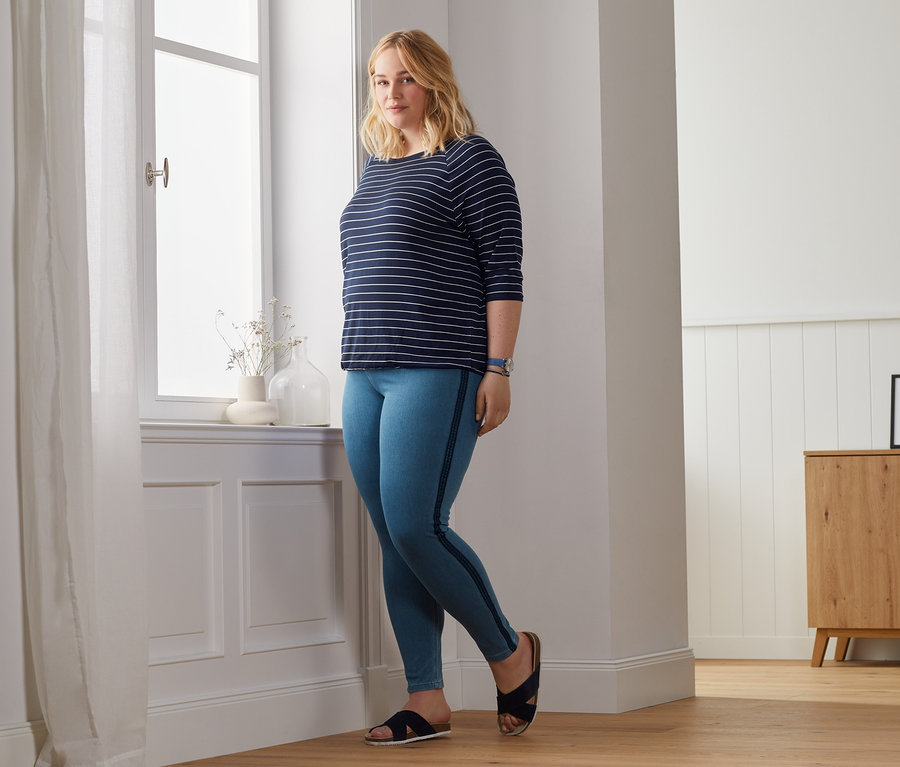 Egy szőke nő pózol az ablaknál denevérujjú felsőben és jeggingsben.