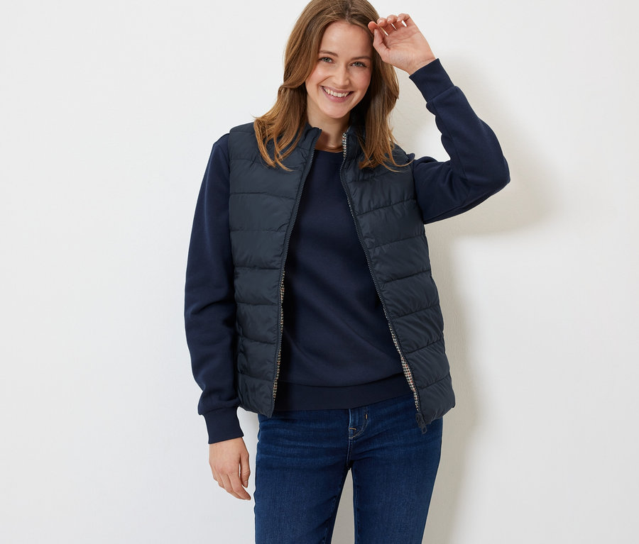 Egy mosolygó nő sötétkék steppelt mellényben, pulóverben és farmerben pózol.