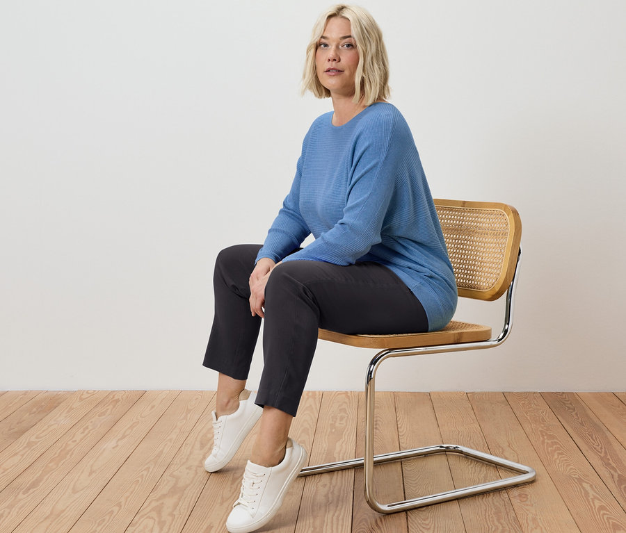 Egy szőke nő ül egy széken, sztreccsnadrágot és egy kék, finom kötésű pulóvert visel.