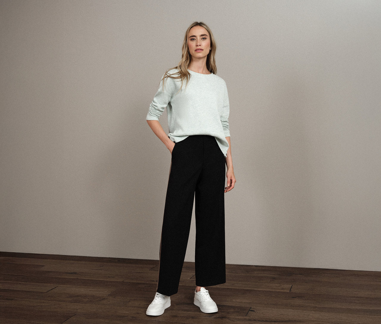 Nő áll zsebre tett kézzel, Street One bordás mintás pólót, fekete nadrágot és fehér tornacipőt visel.