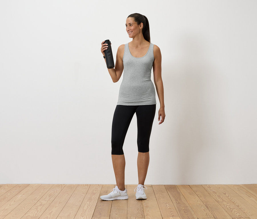 Nő tart egy vizespalackot, szürke trikót, fekete leggingset és fehér tornacipőt visel.