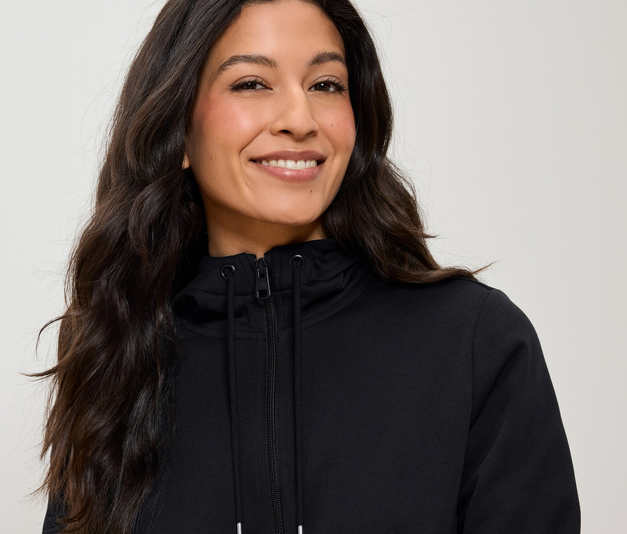 Portré egy hosszú, barna hajú nőről, aki fekete, cipzáros kapucnis pulóvert visel.