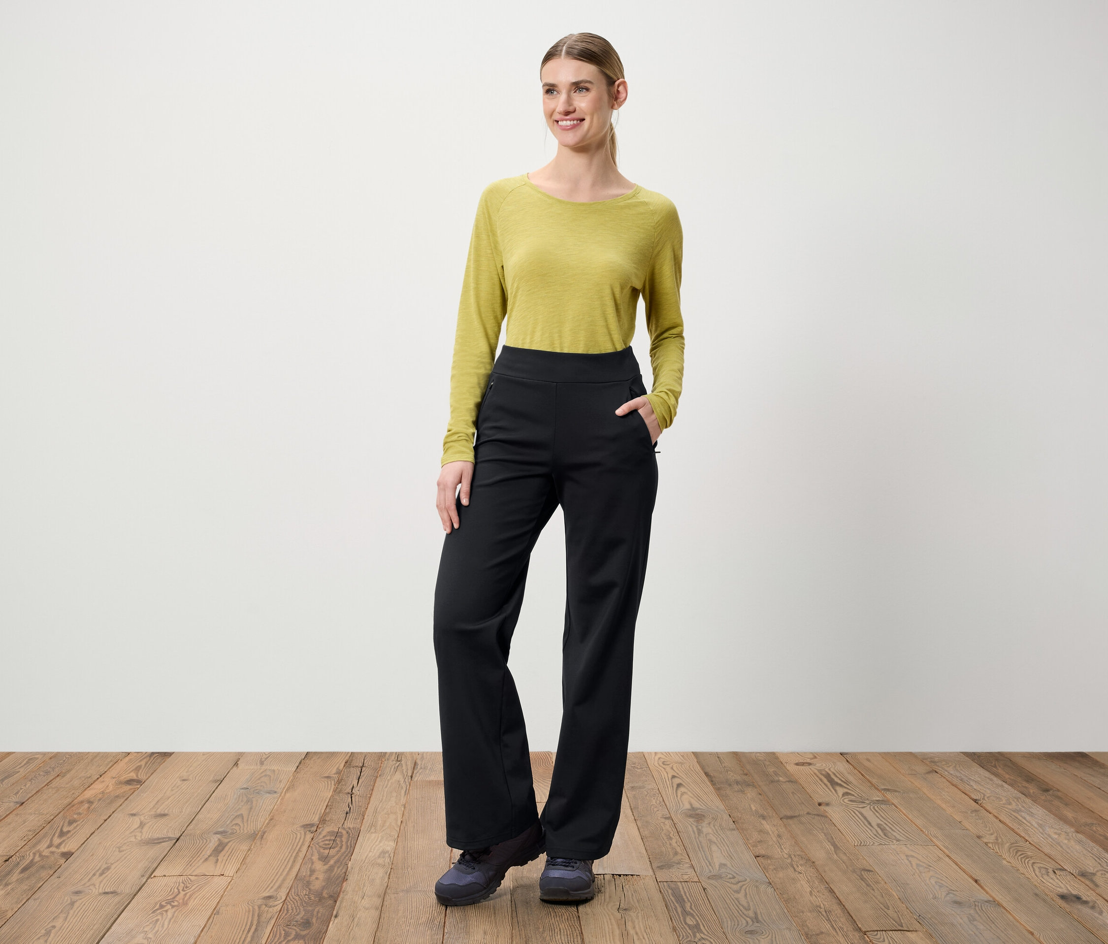 Egy nő áll egy fa padlón. Zöld, hosszú ujjú blúzt és fekete nadrágot visel.