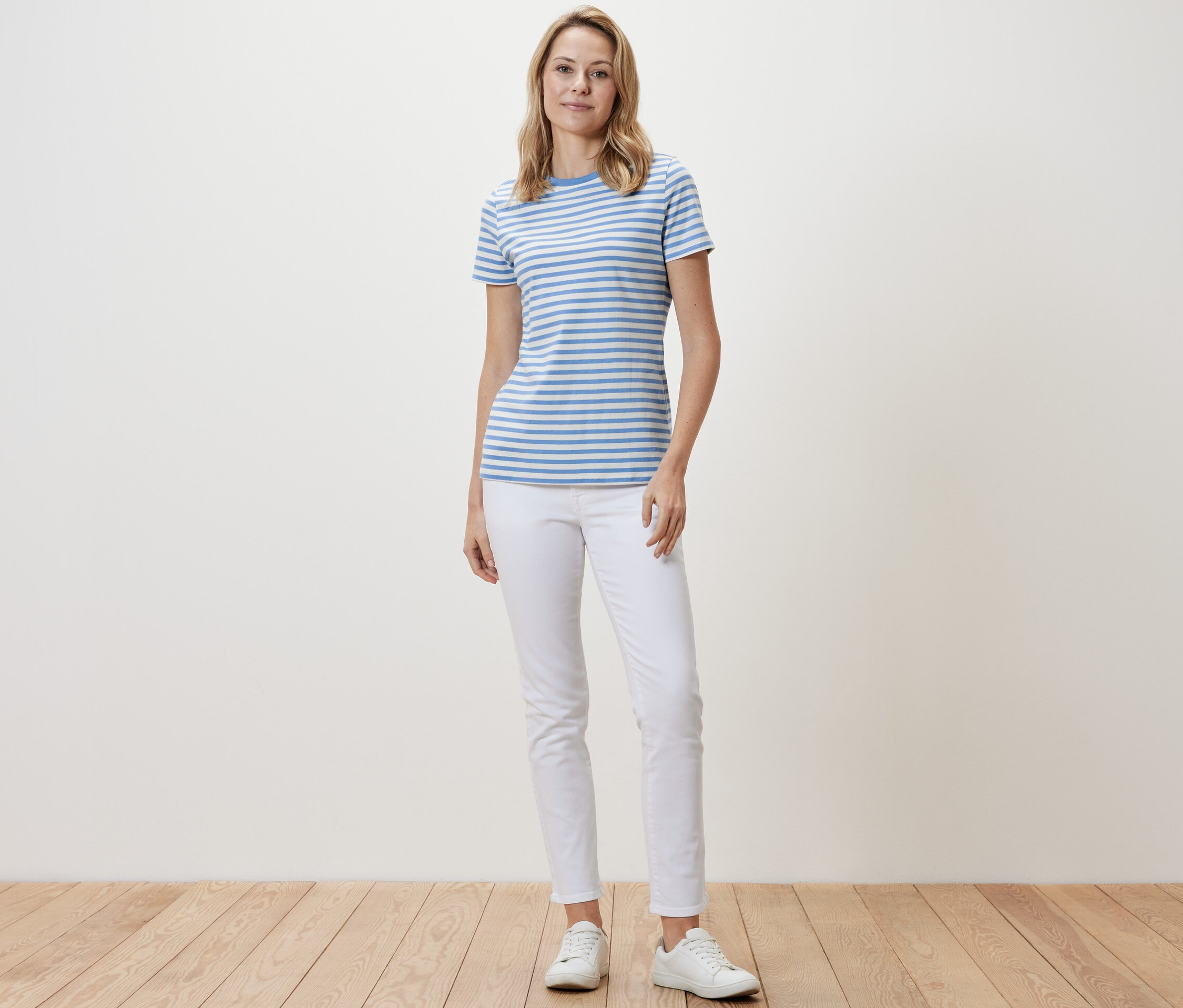 Egy nő áll, és csíkos, világoskék/fehér női pólót, fehér nadrágot és fehér tornacipőt visel.