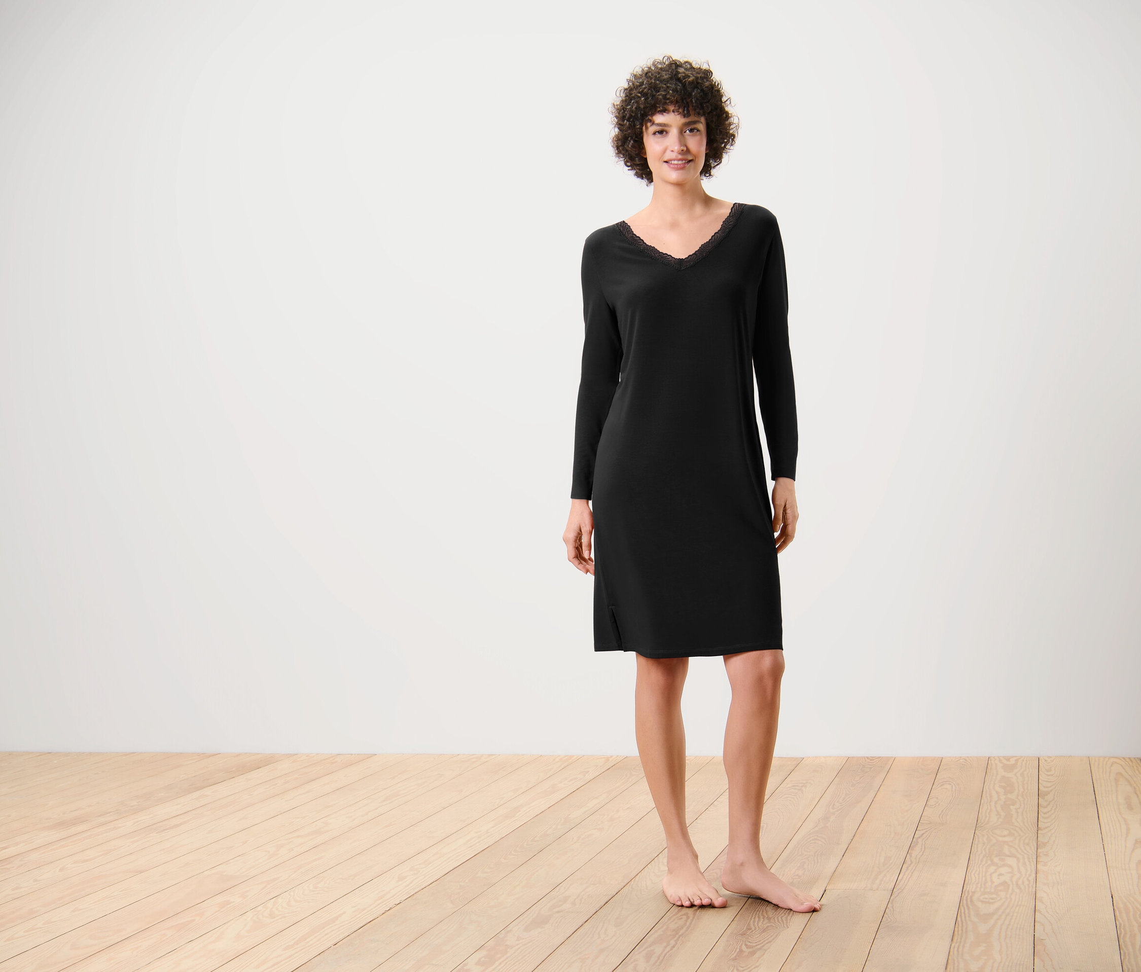 Egy nő áll mezítláb a fapadlón, és egy fekete, hosszú ujjú ruhát visel.