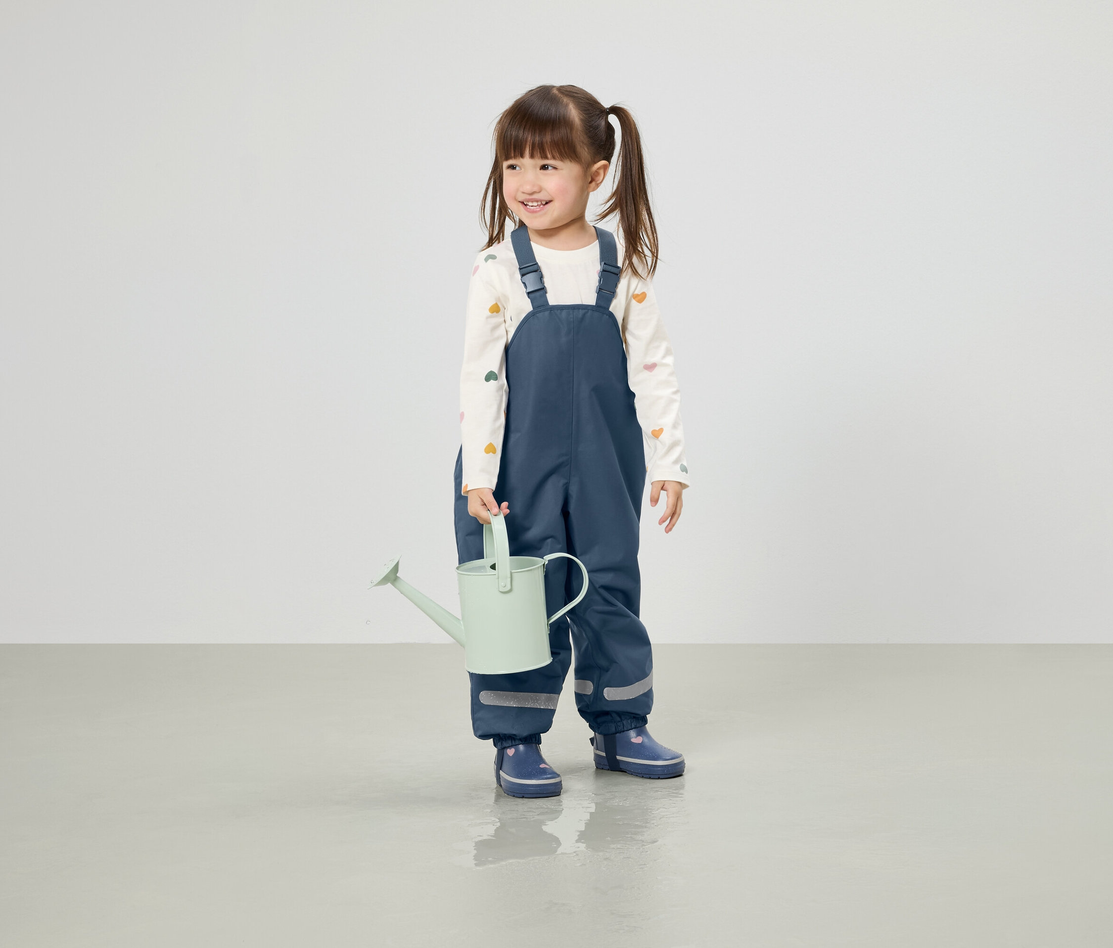 Mosolygó kislány copfokkal világoszöld locsolókannát tart, kék nadrágba és gumicsizmába öltözve.