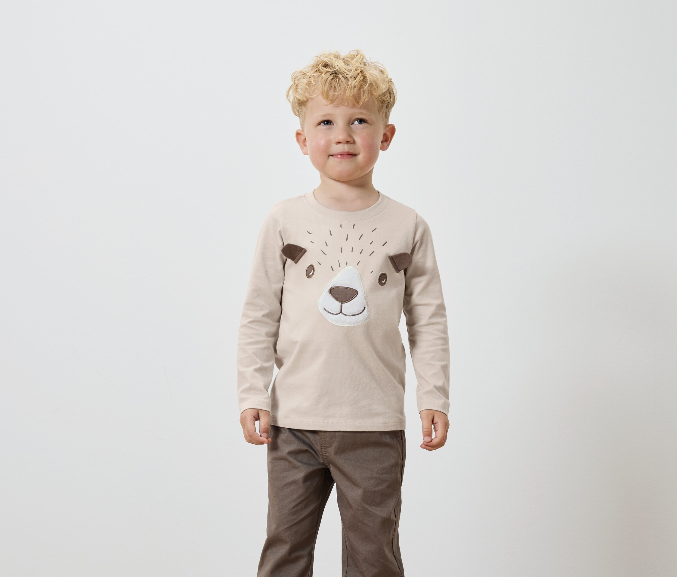 Egy szőke, göndör hajú fiú bézs színű, hosszú ujjú inget visel, barna medvearc részletekkel, valamint barna nadrágot.