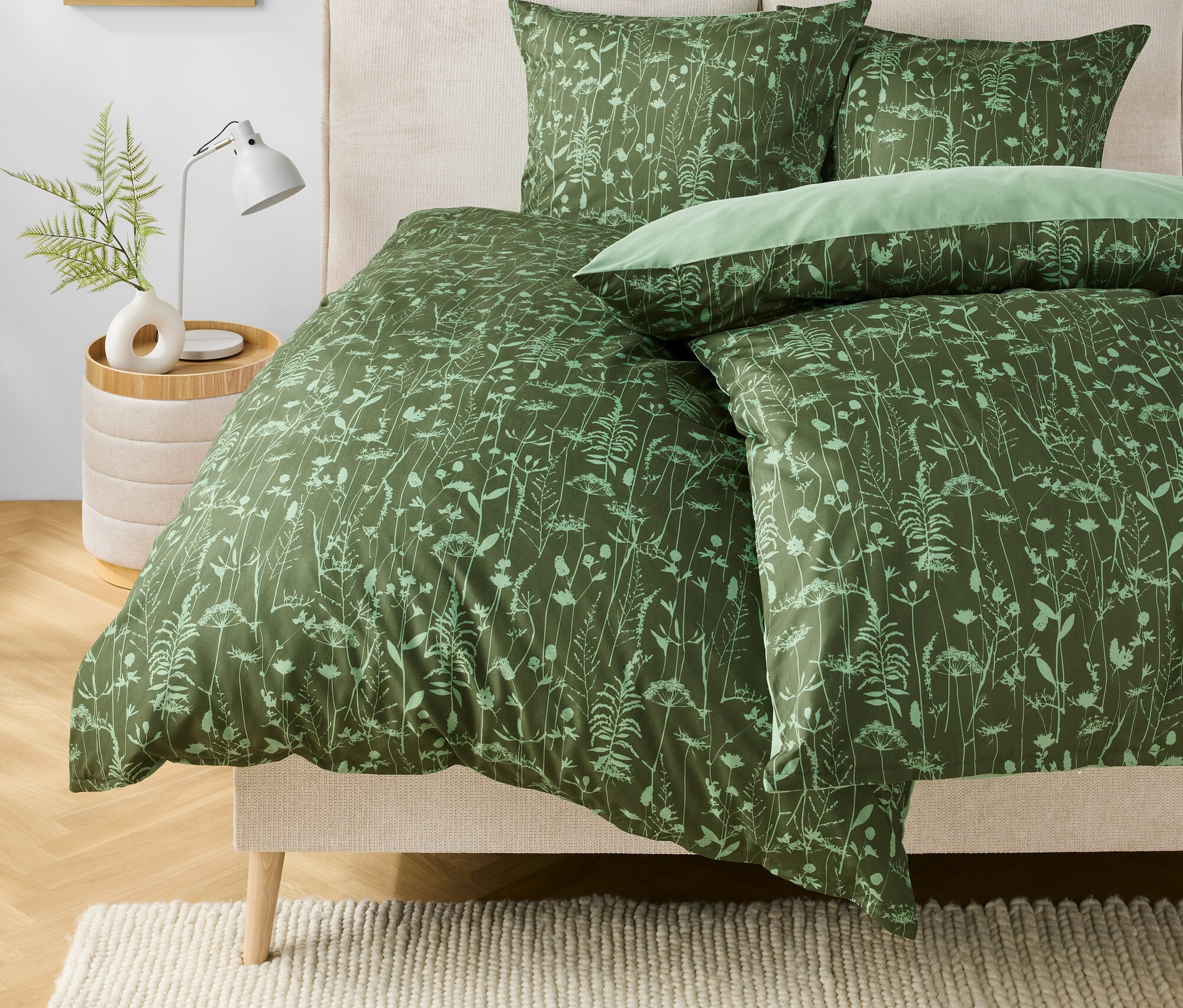 Zöld virágmintás ágynemű egy ágyon. Az éjjeliszekrényen egy lámpa és egy váza áll egy növényel.