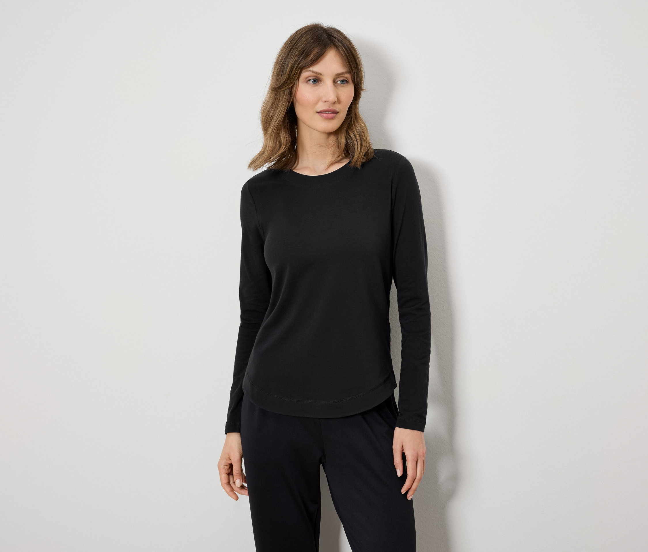 Egy nő fekete hosszú ujjúban és fekete nadrágban áll egy fehér fal előtt.