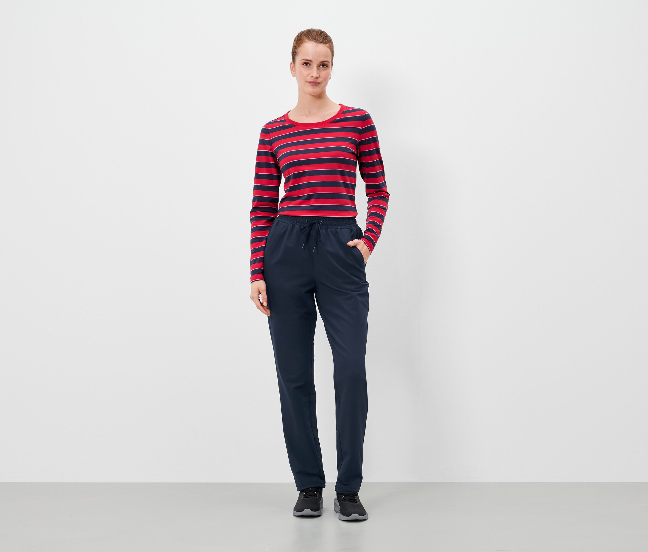 Egy nő áll, kezét a sötétkék nadrágja zsebében tartja, és piros-kék csíkos felsőt visel.