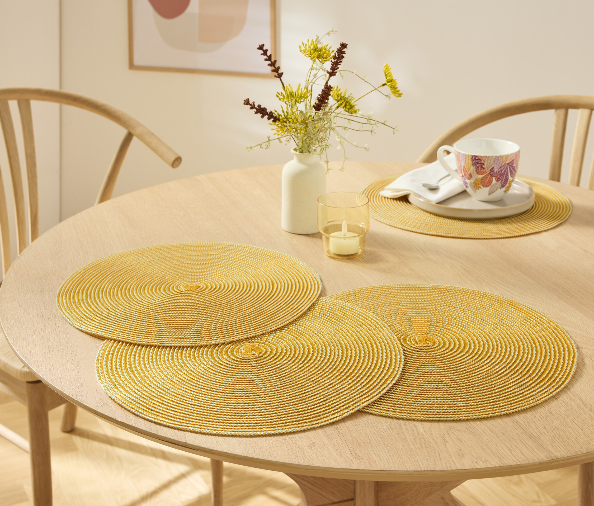 Egy világos fa asztalon három sárga, kör alakú tányéralátét, egy váza virágokkal, egy gyertya és egy csésze alátéttel.
