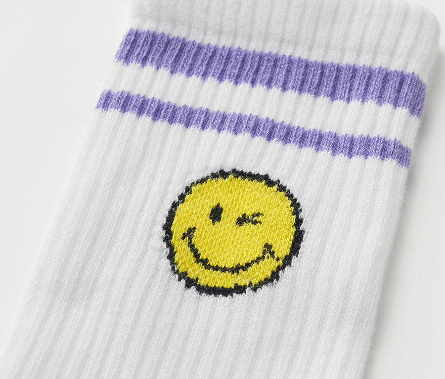Egy fehér gyerek tenisz zokni részlete lila csíkokkal és sárga mosolygós arccal az 5 pár SMILEYWORLD® gyerek sportzokni szettből.
