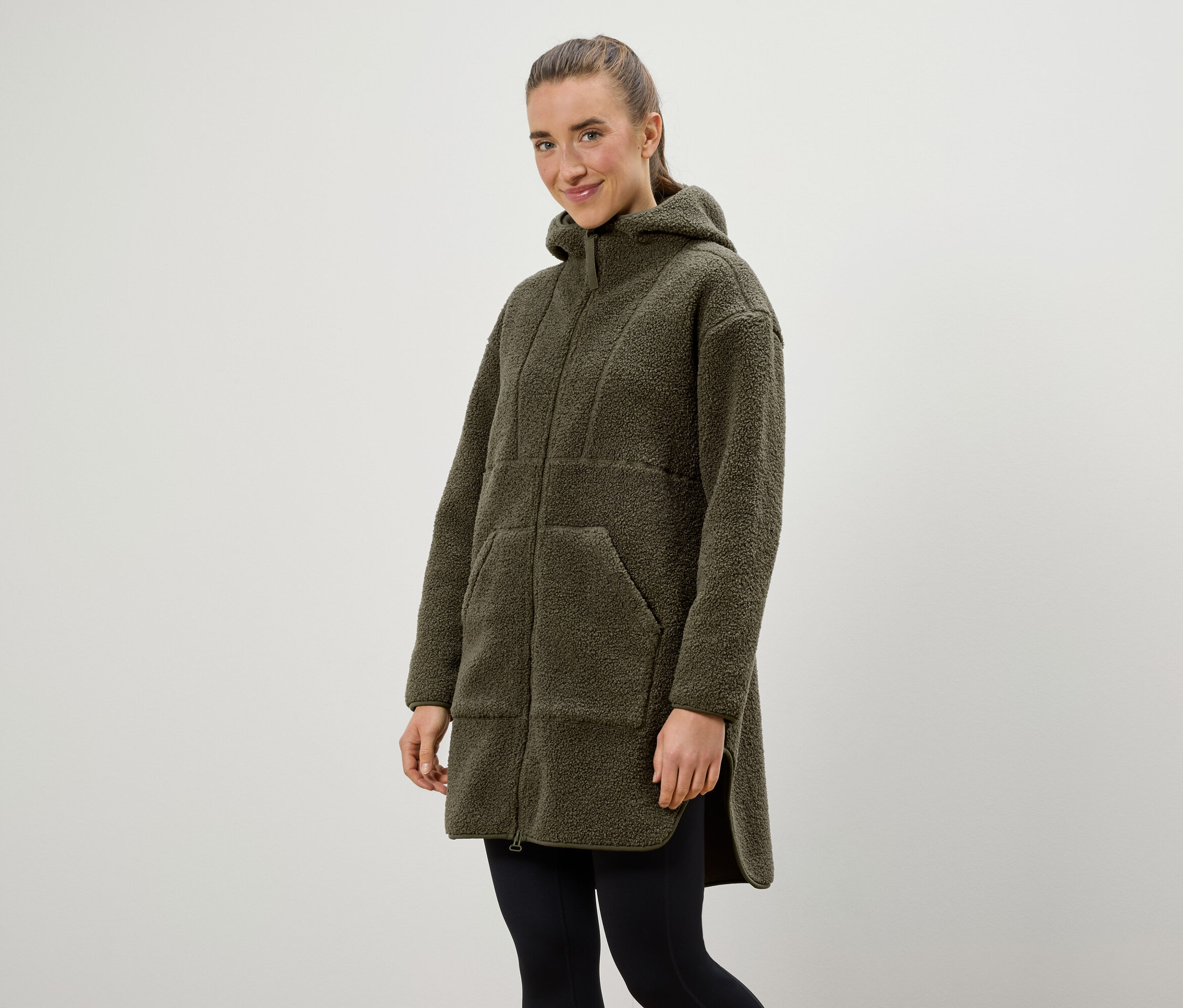 Nő visel olívazöld kapucnis kabátot és fekete leggingset.